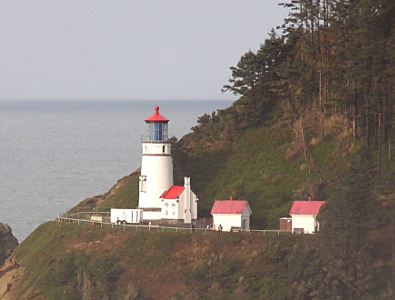 [A white lighthouse with a red cap with a house-like building which appears attached to the inland side. The house is also white with a red roof and has a white chimney on the inland side. Two other white buildings with red roofs are in the same area with a fence marking the edge of the cliff.]